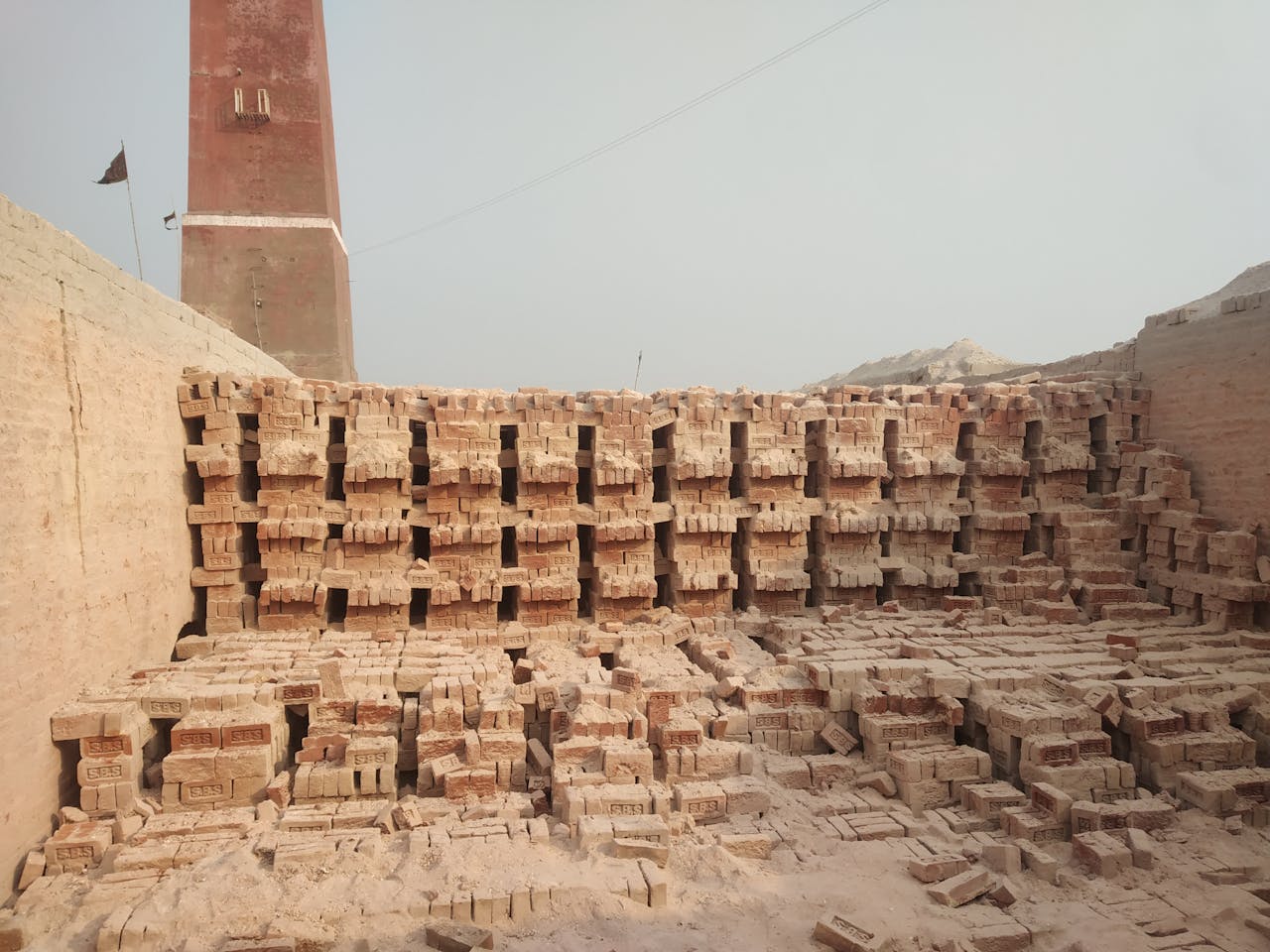 A large stack of clay bricks near a kiln tower in a New Delhi brick factory yard.